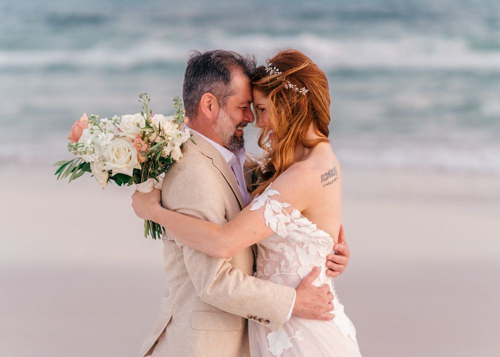 A romantic beach wedding couple embraces by the ocean waves while holding a white floral bouquet.
