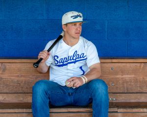 Teen boy in Sapulpa baseball jersey holding bat and ball sitting in dugout bench.