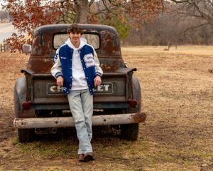 Teen boy in letterman jacket leaning against a vintage rusty Chevrolet truck in a rural outdoor setting.