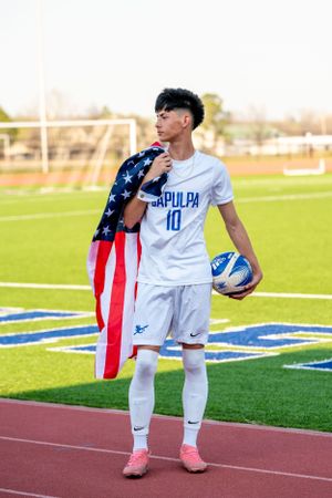 High school senior soccer player in Sapulpa number 10 jersey holding a ball and American flag on the field.