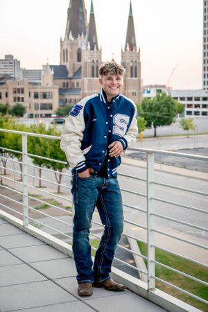 Smiling teen in varsity jacket leaning on railing with cathedral and city skyline behind him.