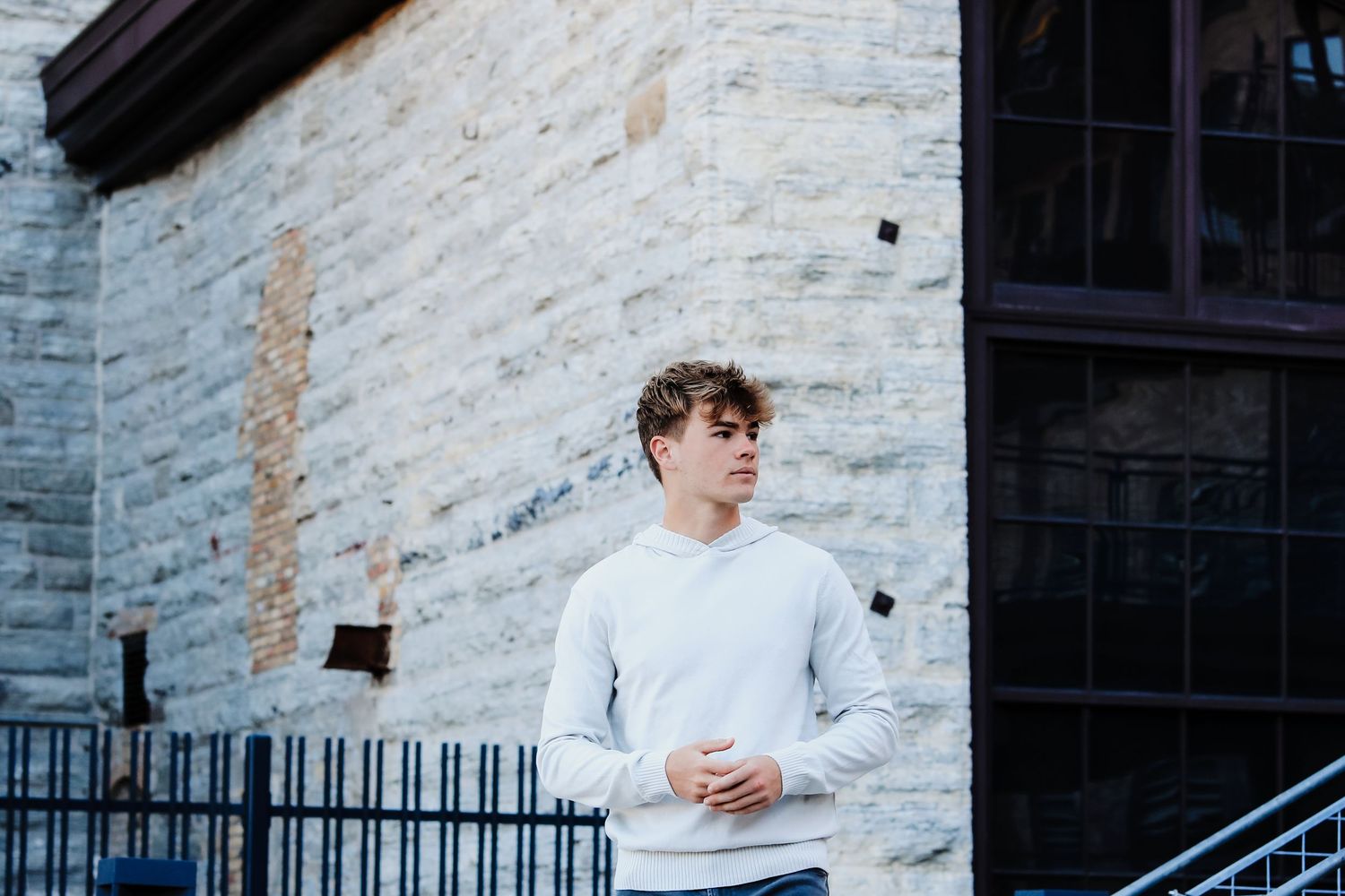 Young man in white sweatshirt standing outdoors near rustic stone building with iron railing.