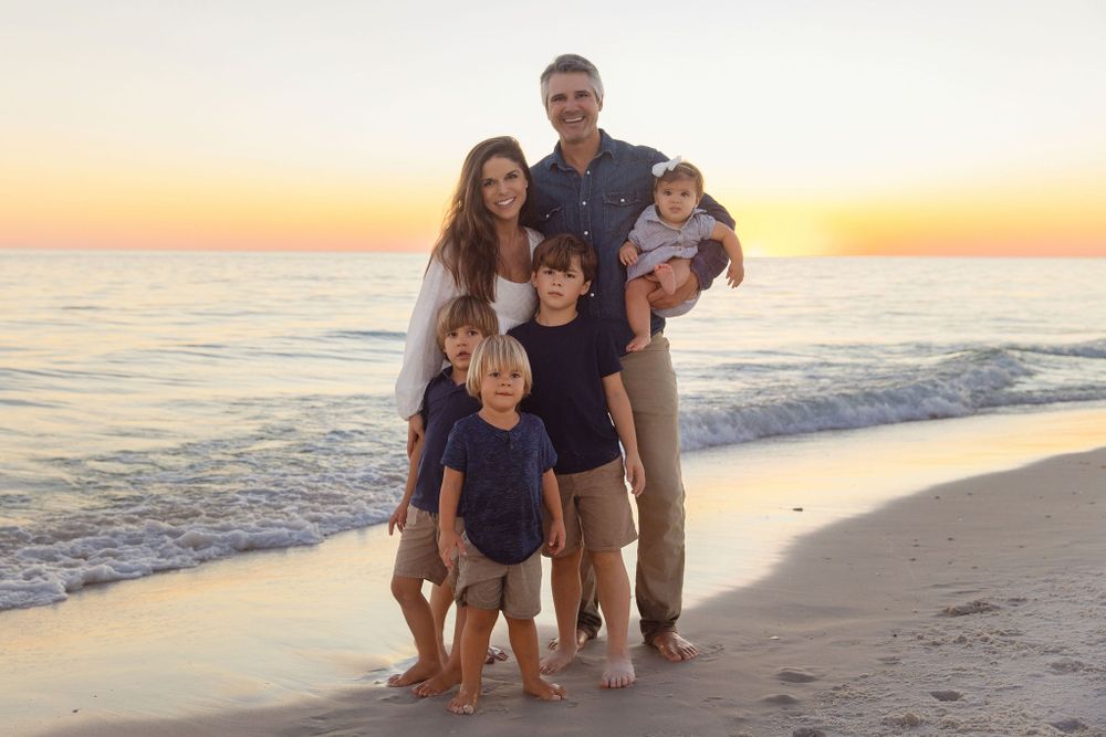 A happy family of six poses together on a sandy beach at sunset with the ocean waves in the background.