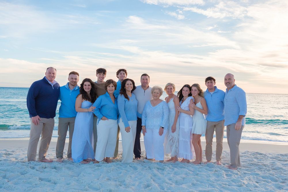 A group in coordinated blue and white outfits poses together on a white sandy beach at sunset.