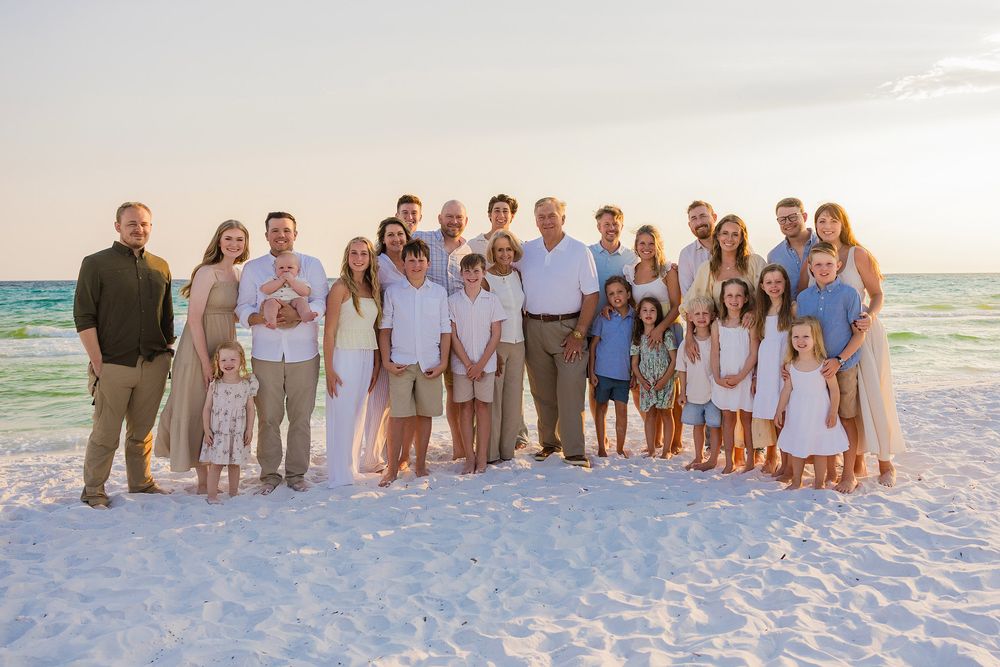 A group of people in coordinated white and blue outfits pose together on a sunny beach at sunset.