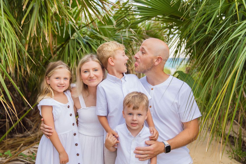 A family wearing coordinated white outfits poses together outdoors against tropical palm plants.