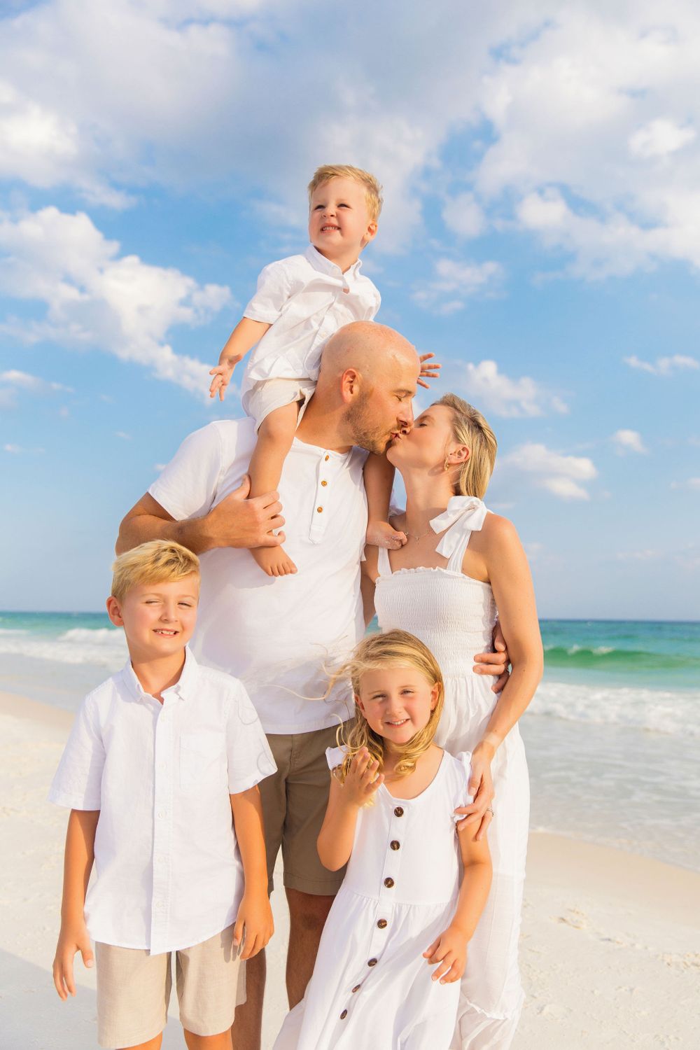 A family in white clothing poses together on a sunny beach with blue ocean waves and fluffy clouds in the background.