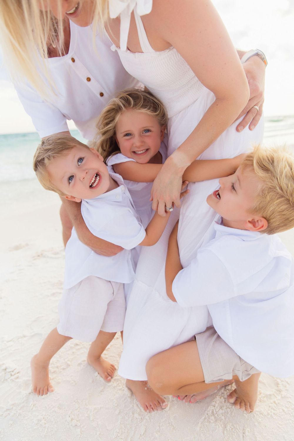 Happy family wearing all white clothing embrace and laugh together on a bright sandy beach.