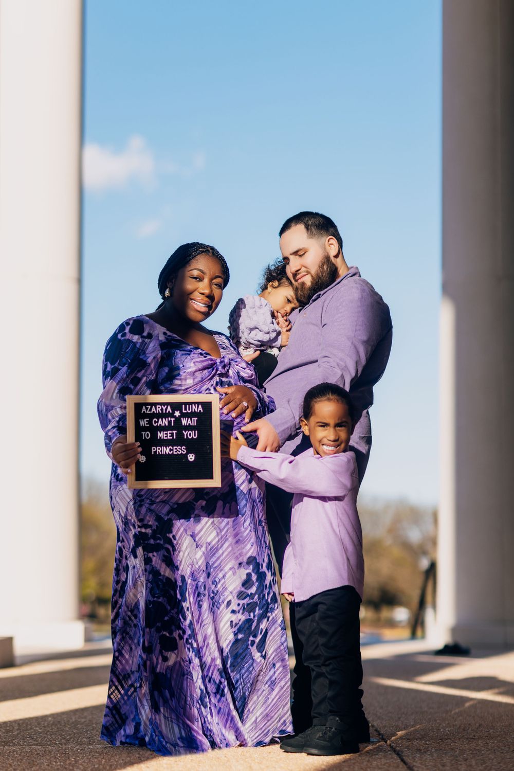 A family poses outdoors with a letter board sign in coordinating purple outfits.