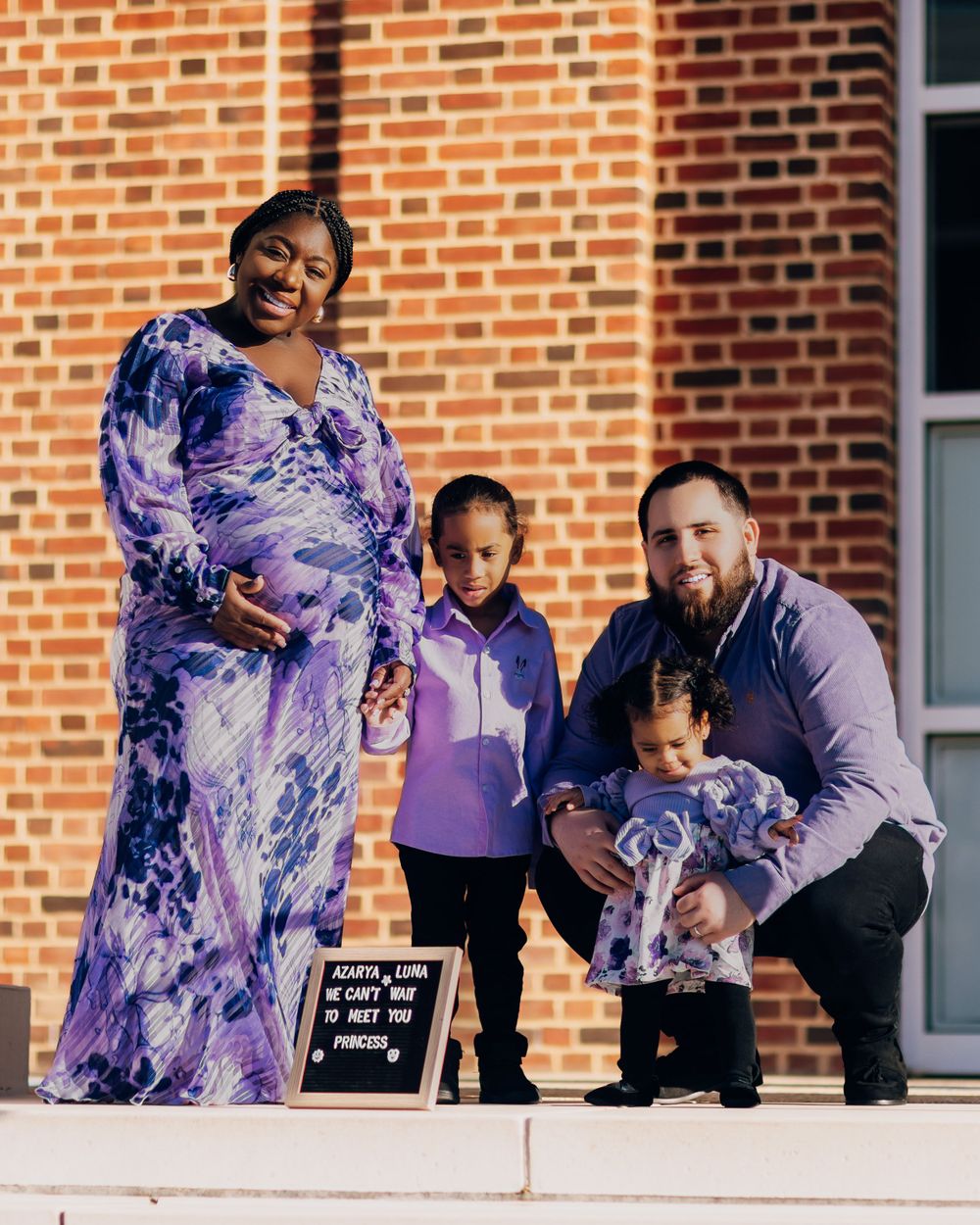 A family group poses on steps in front of a brick building wearing coordinated purple outfits.