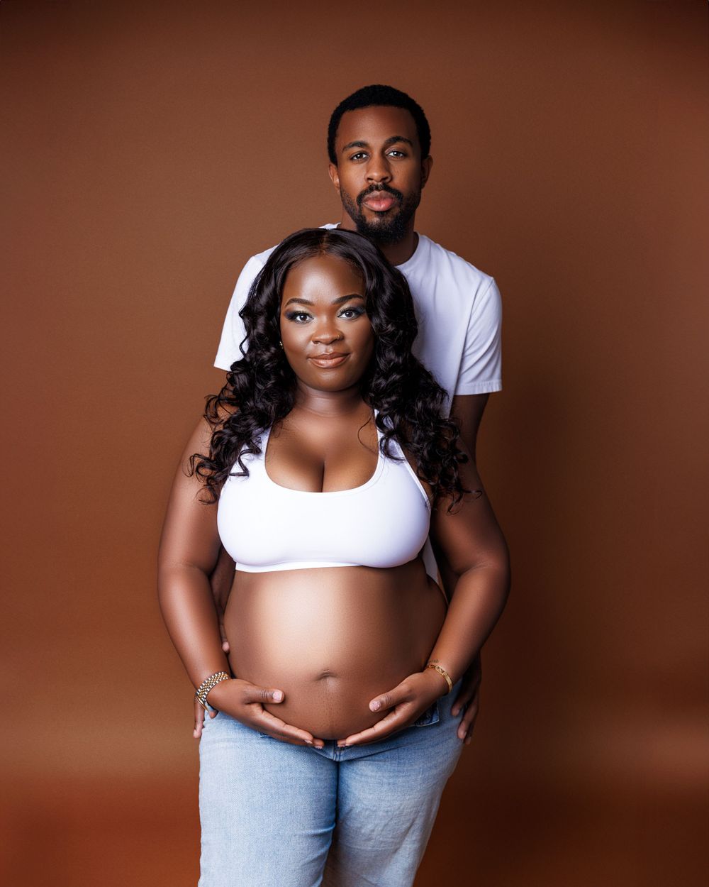 A maternity portrait of a couple in white tops against a brown background.