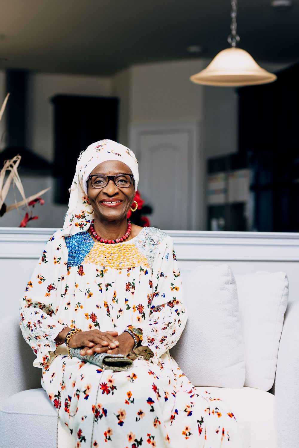 Portrait of woman in floral dress and white headwrap sitting on white couch.