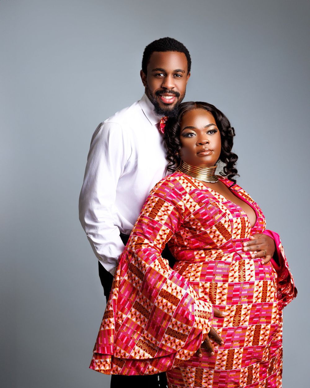 A couple poses for a maternity photo, with the woman wearing a vibrant red and yellow patterned dress.