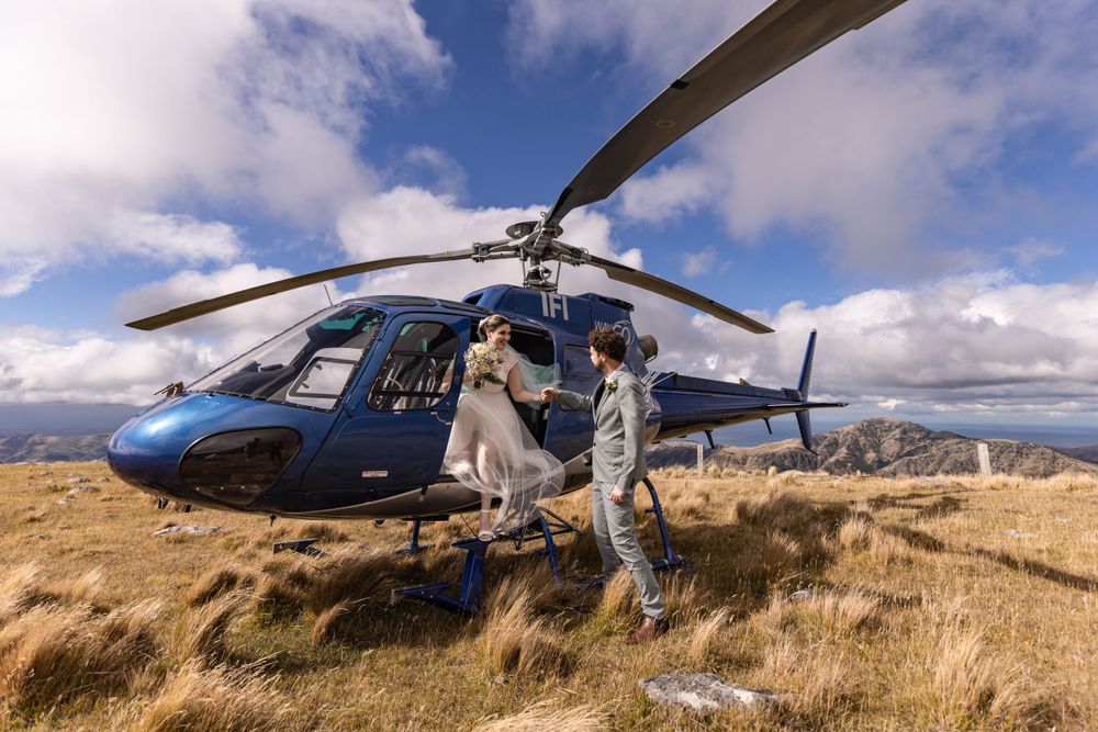 A blue helicopter sits on a grassy mountainous ridge while passengers prepare to disembark on a sunny day.