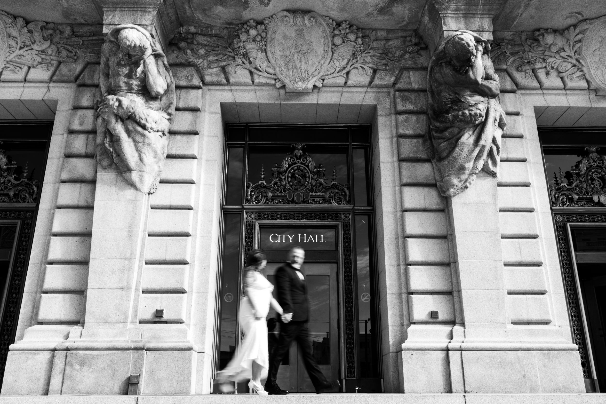 Black and white photo of a wedding couple exiting a building with classical stone carvings.