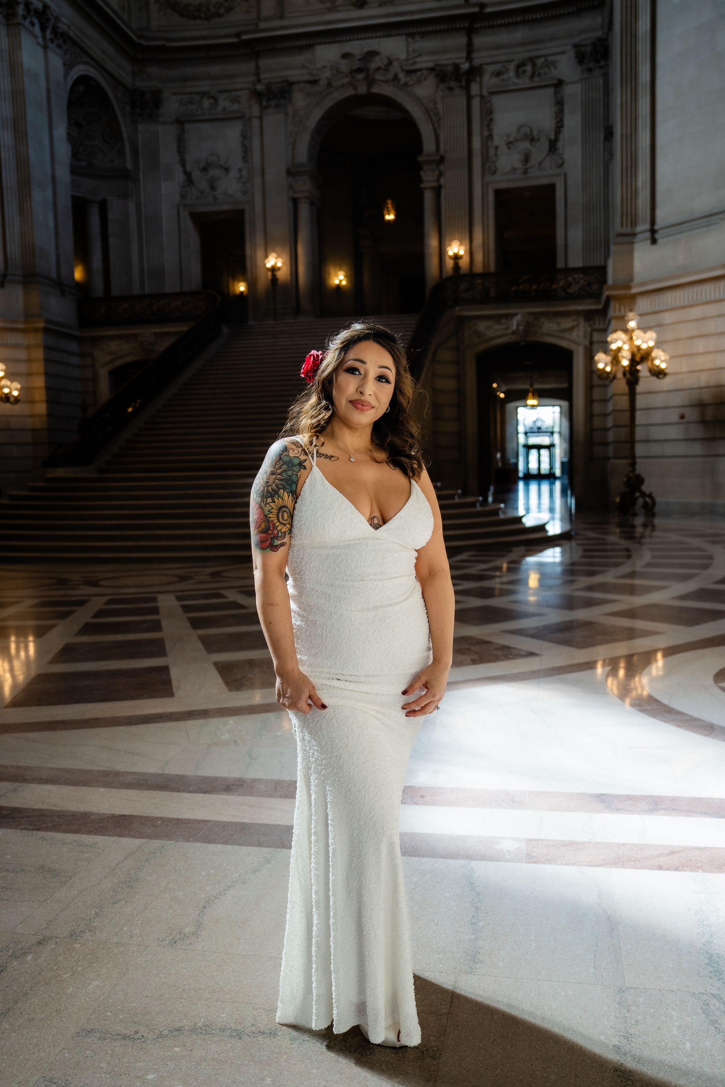 Bride in fitted white dress stands in marble hall with dramatic natural lighting.