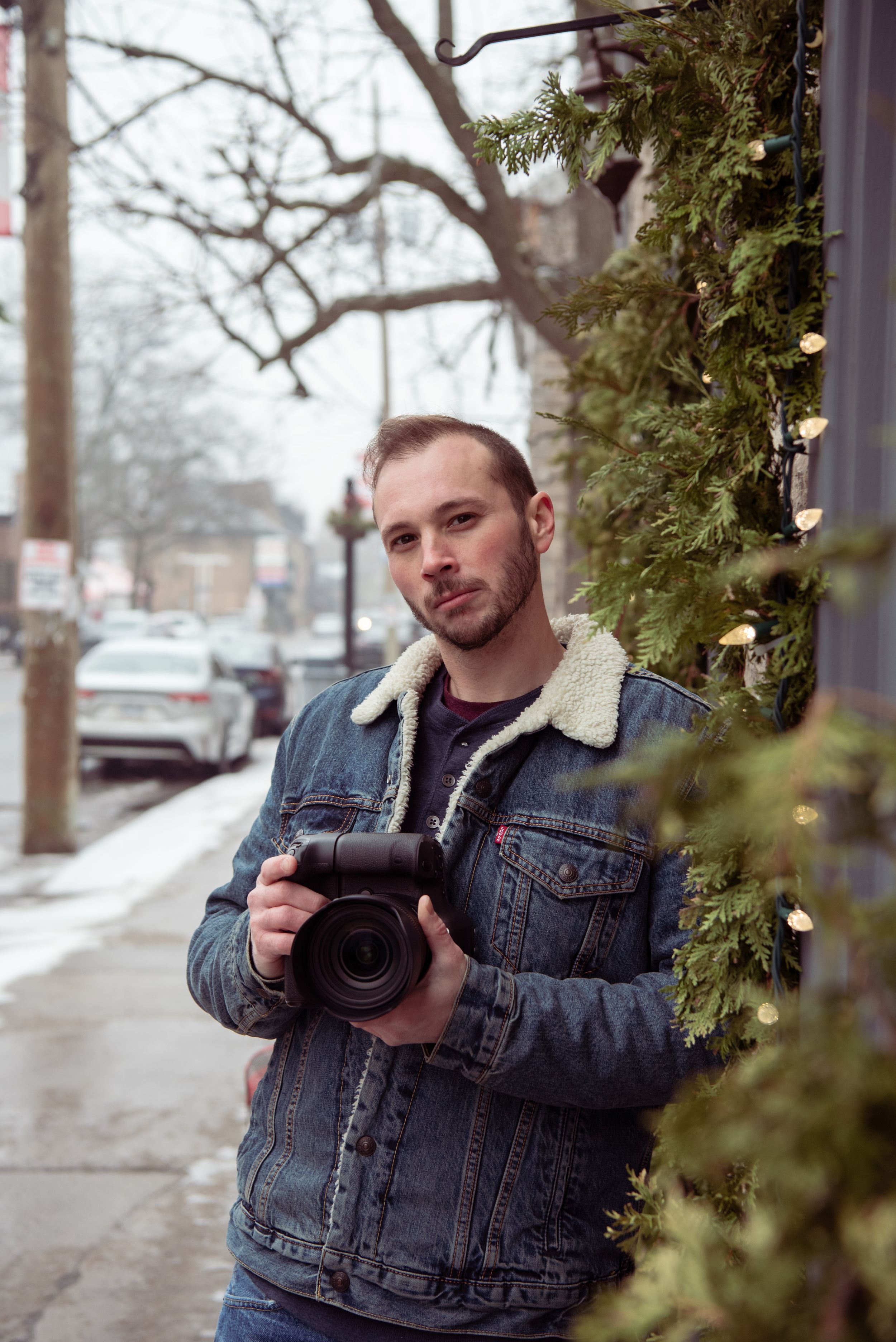 A photographer in a sherpa-lined denim jacket holds a professional camera on a winter street.