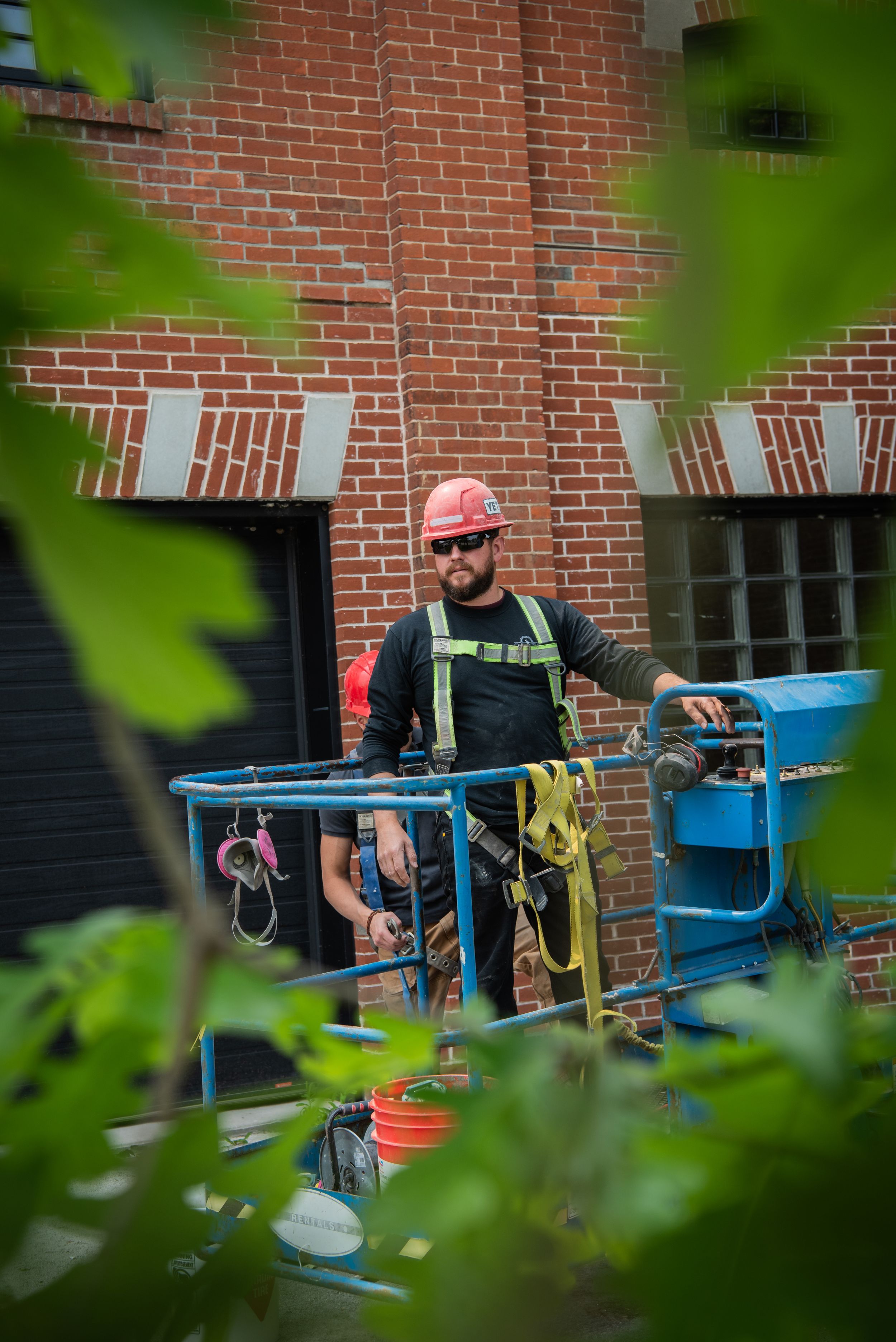 A person in a red hat works with equipment on a table against a brick wall, framed by green foliage in the foreground.