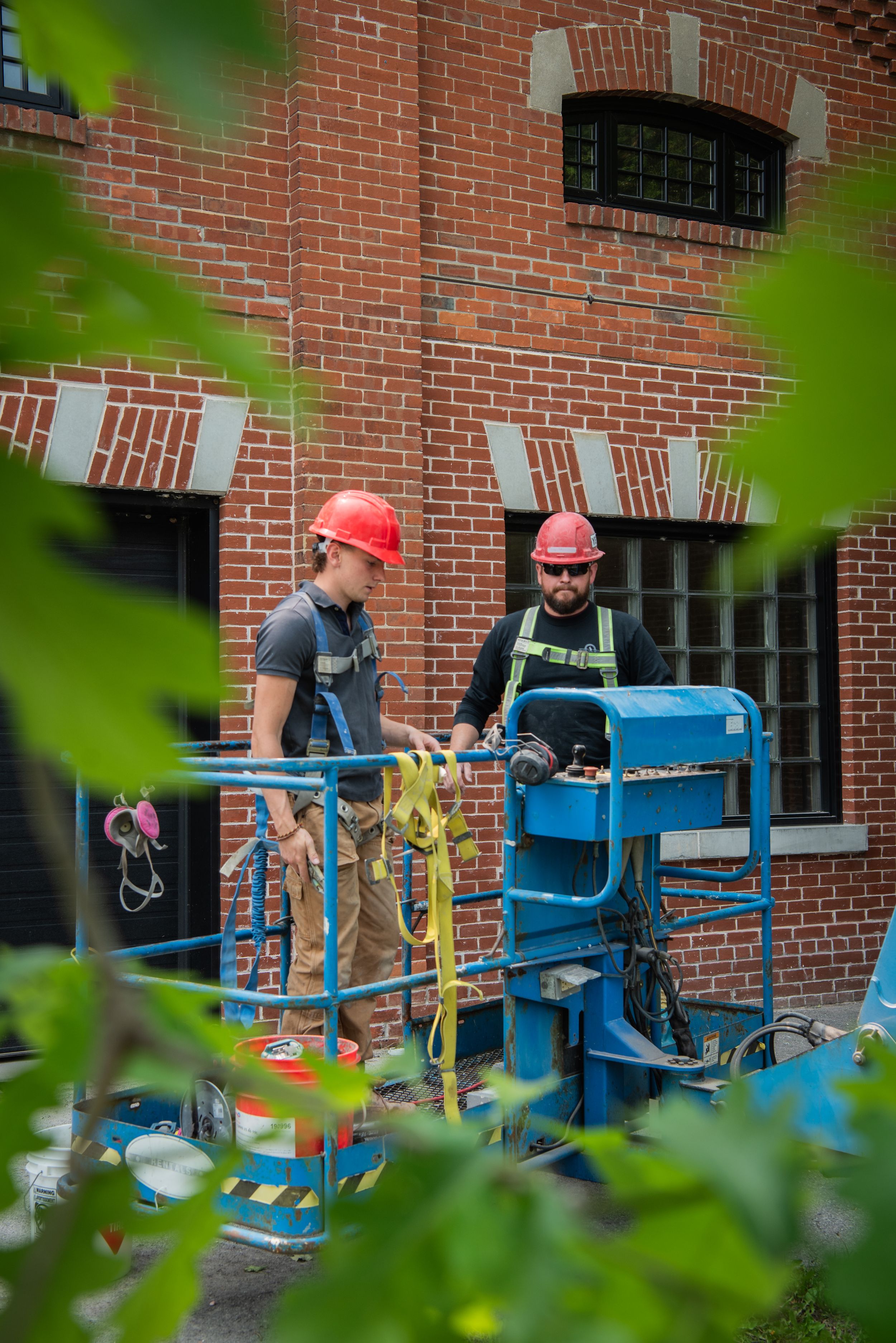 Workers on elevated lift platforms perform maintenance work on the exterior of a brick building.