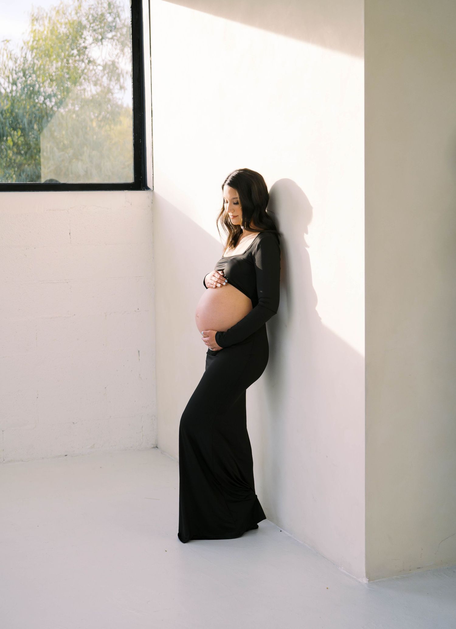 Elegant maternity photo of woman in long black dress posing by window in minimalist white room with natural light.