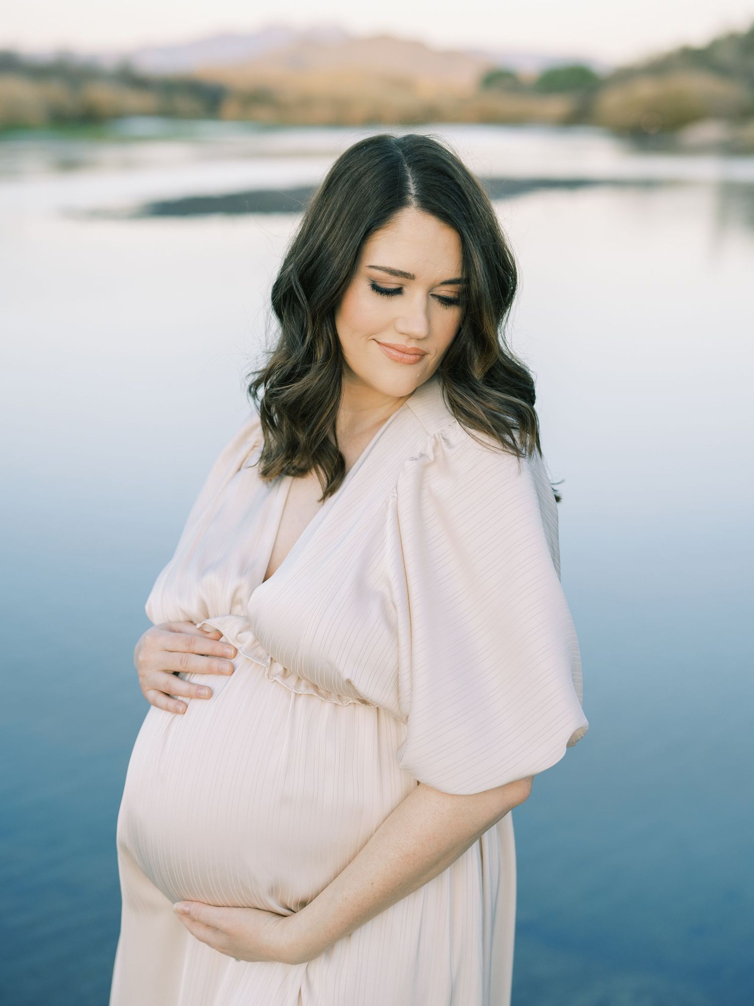 A maternity portrait by a serene lake with mountains in the background.