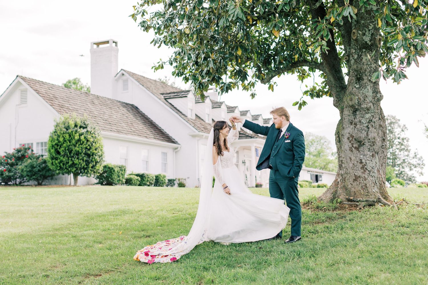 Groom twirls bride in white wedding gown on green lawn beside a white barn and large tree.