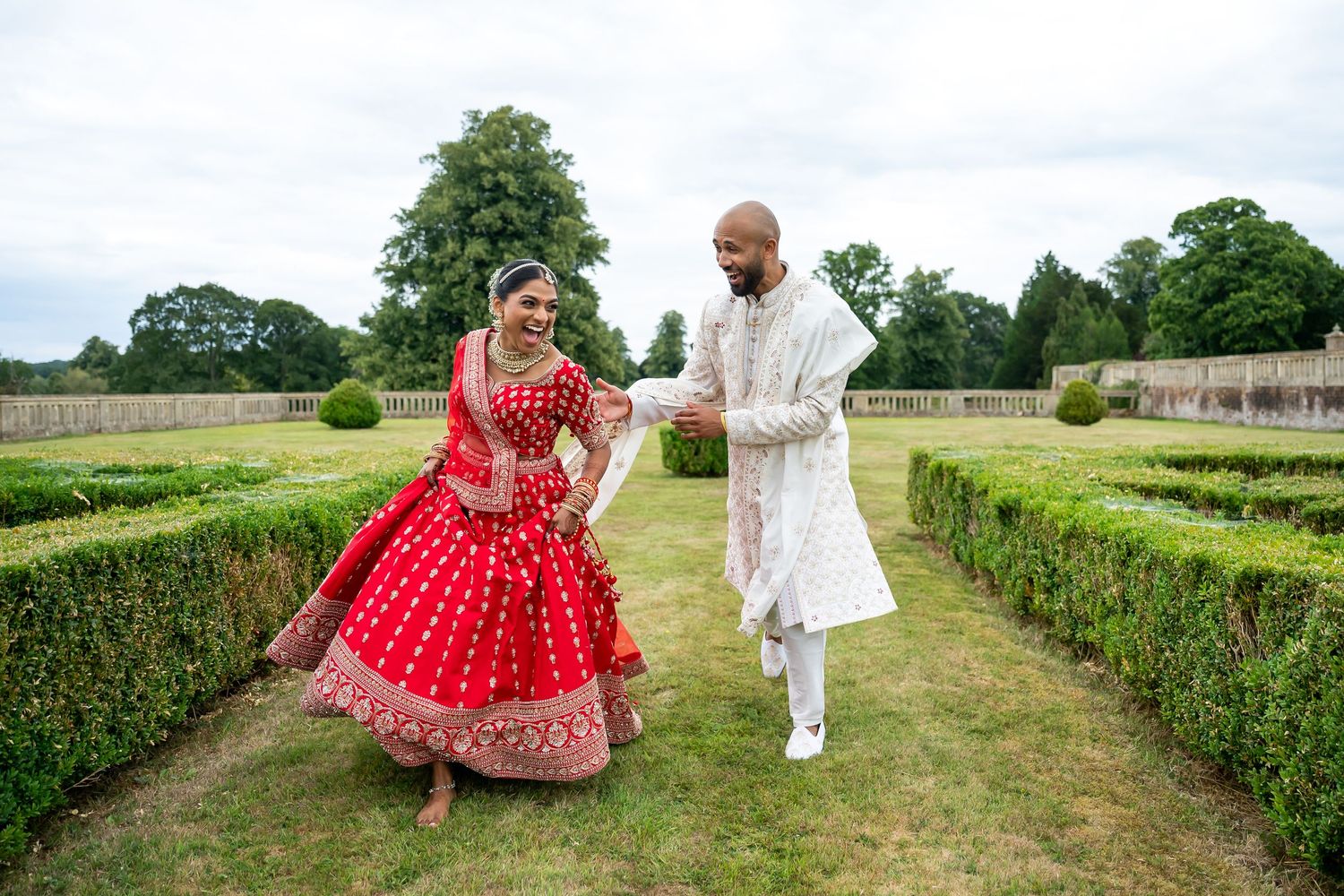 Indian bride in red lehenga and groom in white sherwani holding hands in a lush outdoor wedding at North Mymms Park during wedding ceremony.