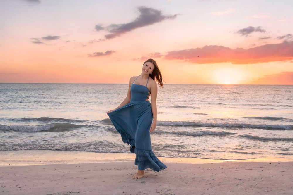 Woman in flowing blue dress poses on sandy beach during vibrant sunset with ocean waves in background.
