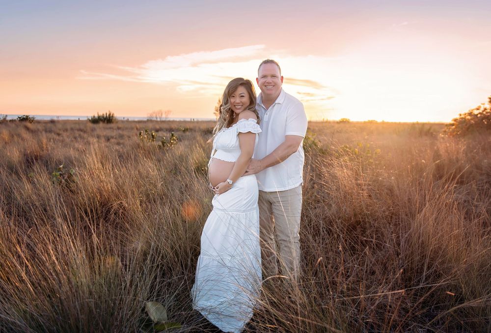 Couple poses in golden field at sunset, woman in white lace maternity dress, man in white shirt, warm sky backdrop.