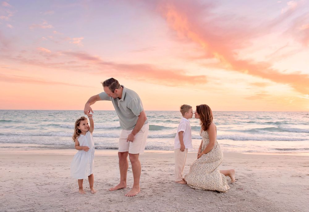 Family of four dancing and playing on a beach at sunset with pink and orange sky in the background.