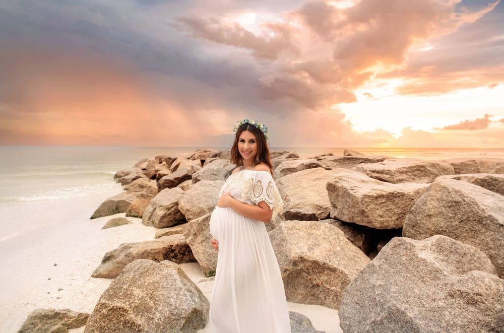 Ethereal maternity photo of a white dress against dramatic sunset sky and rocky beach landscape.