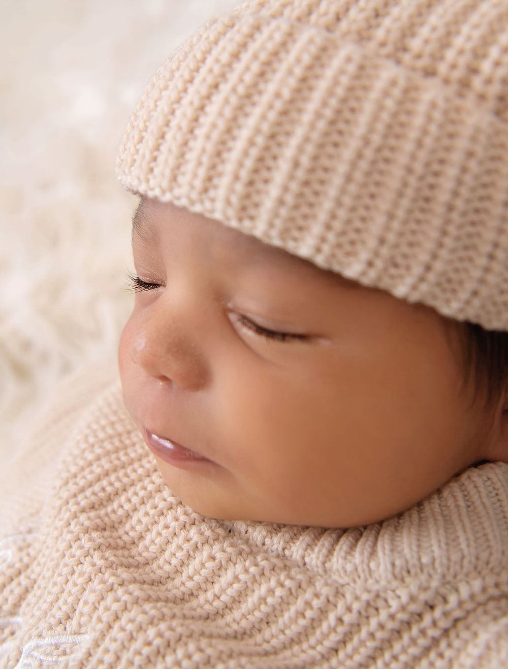 Sleeping newborn baby wearing a cozy beige knit hat and sweater, peacefully resting on a soft cream blanket.