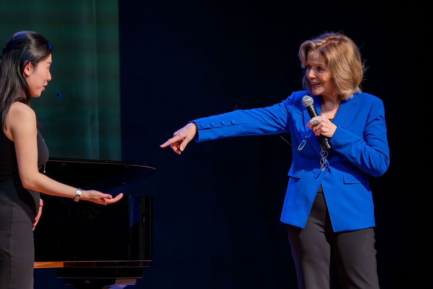 Performance scene with pianist and speaker in blue blazer interacting on dark stage.