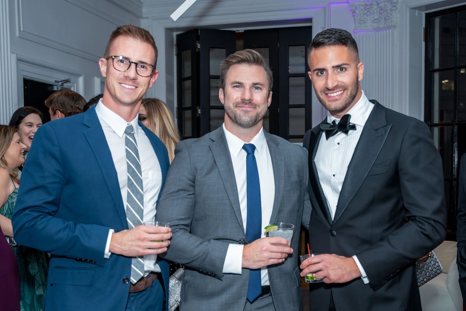 Three men in formal attire smile while holding drinks at an upscale event.