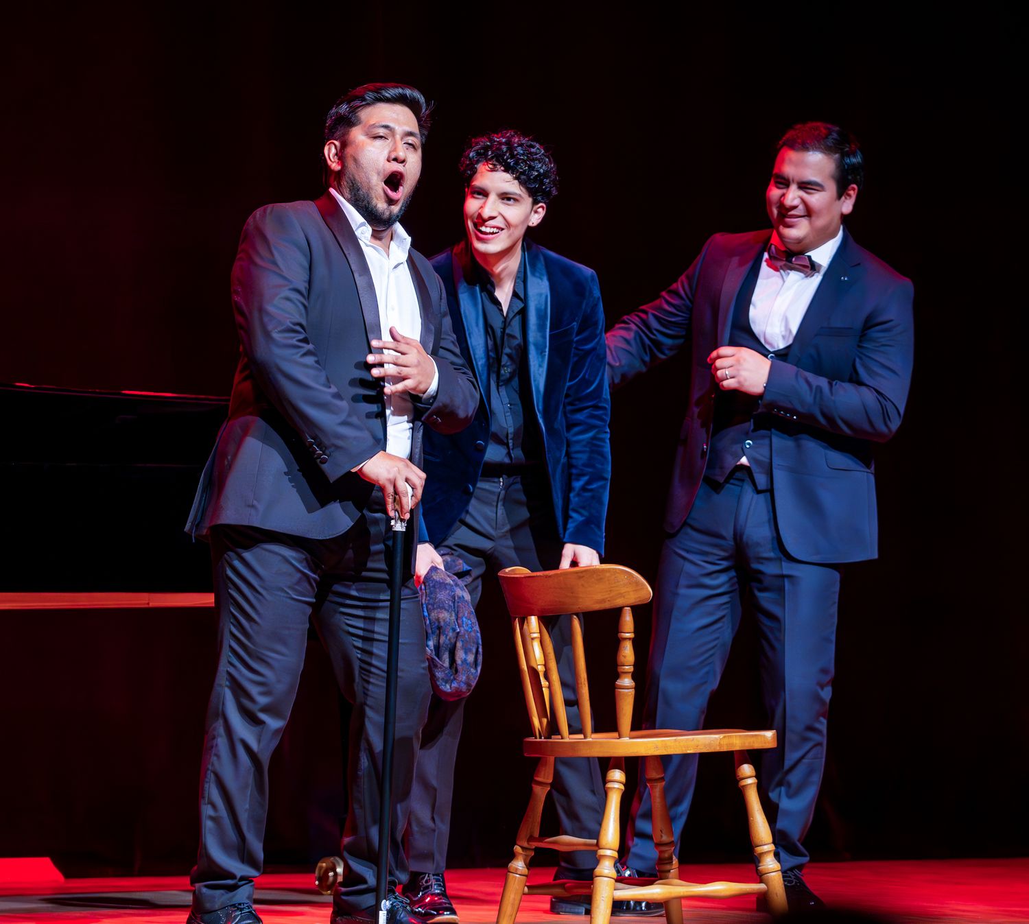 Three performers in suits sing together on stage during a theatrical performance.
