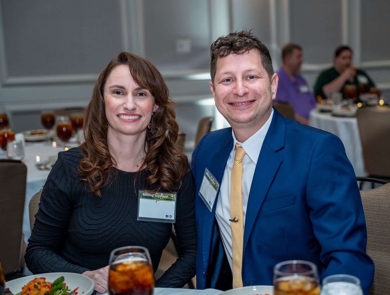 Formal event attendees in business attire seated at dining tables.