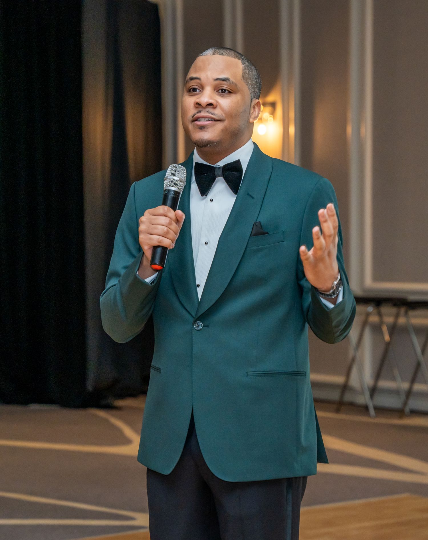 Speaker in teal blazer and bowtie presenting at formal event with microphone.