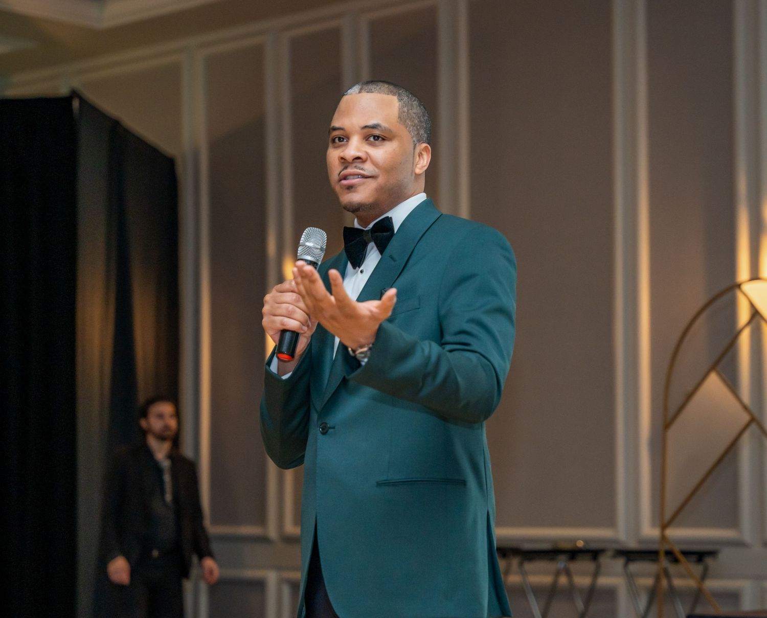 Presenter in emerald green suit jacket speaking on stage at formal gathering.
