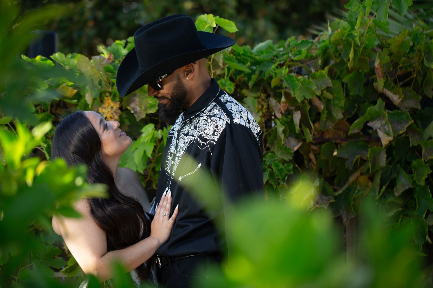 A romantic couple embraces among lush green foliage in a garden setting during a photoshoot.