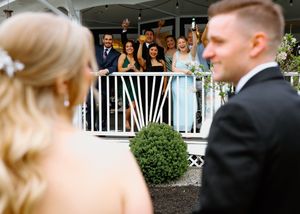 Wedding guests cheer from balcony as bride and groom walk past in foreground.