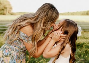 A mother tenderly kisses her young daughter's face outdoors in a green field during a family photo session.