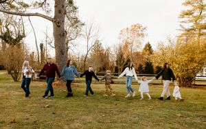 Extended family holds hands walking together across green lawn among autumn trees outdoors.