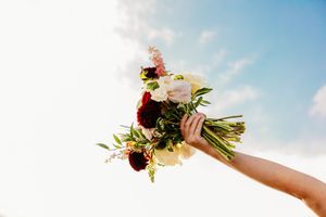 Hand holding a bridal bouquet of white, cream, and burgundy flowers against a bright blue sky.