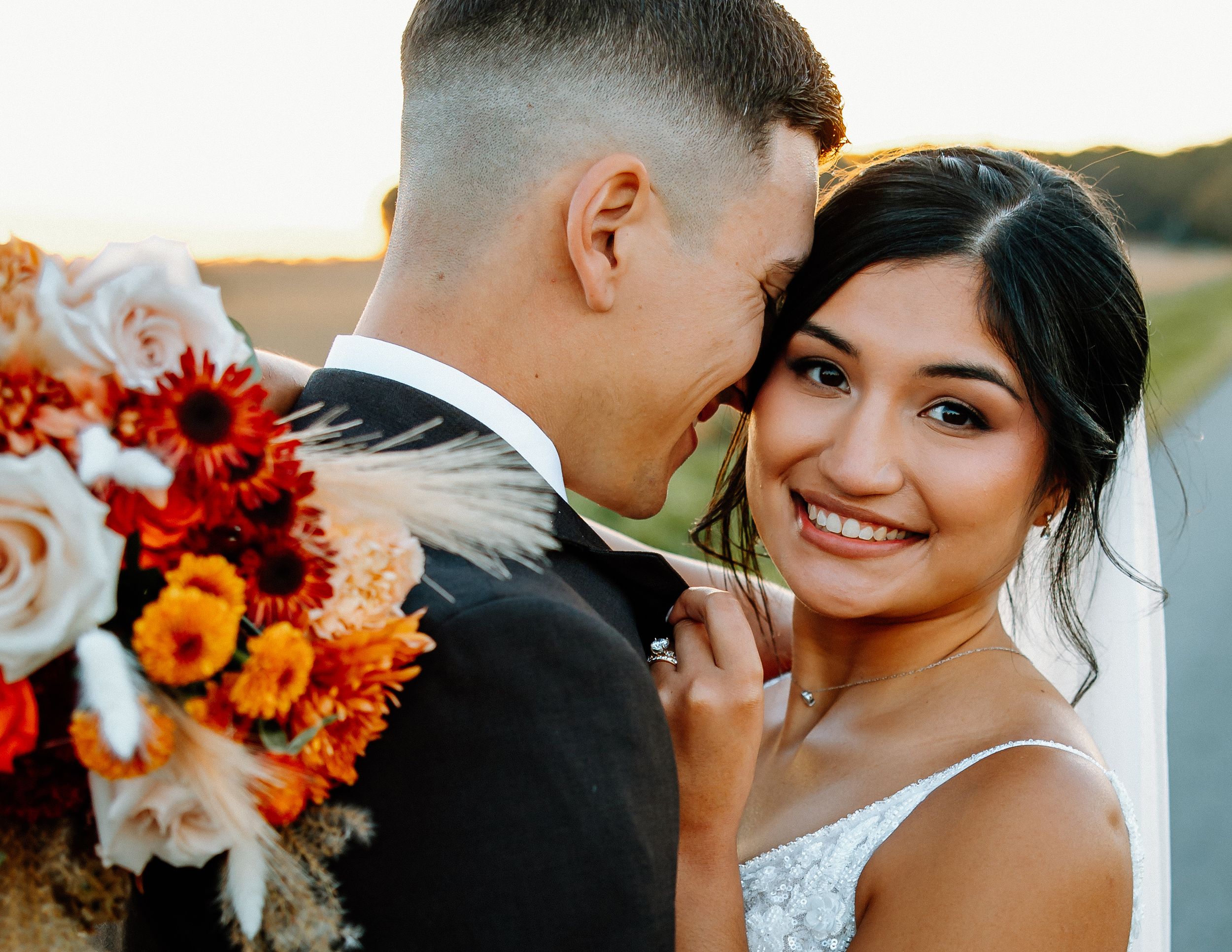 Smiling bride in lace gown holds autumn bouquet of sunflowers and roses with groom at golden hour.
