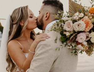 Bride and groom share a kiss outdoors, bride holding lush peach and green floral wedding bouquet.