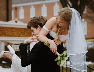 Bride in white veil leans down to embrace groom outside historic church in winter.