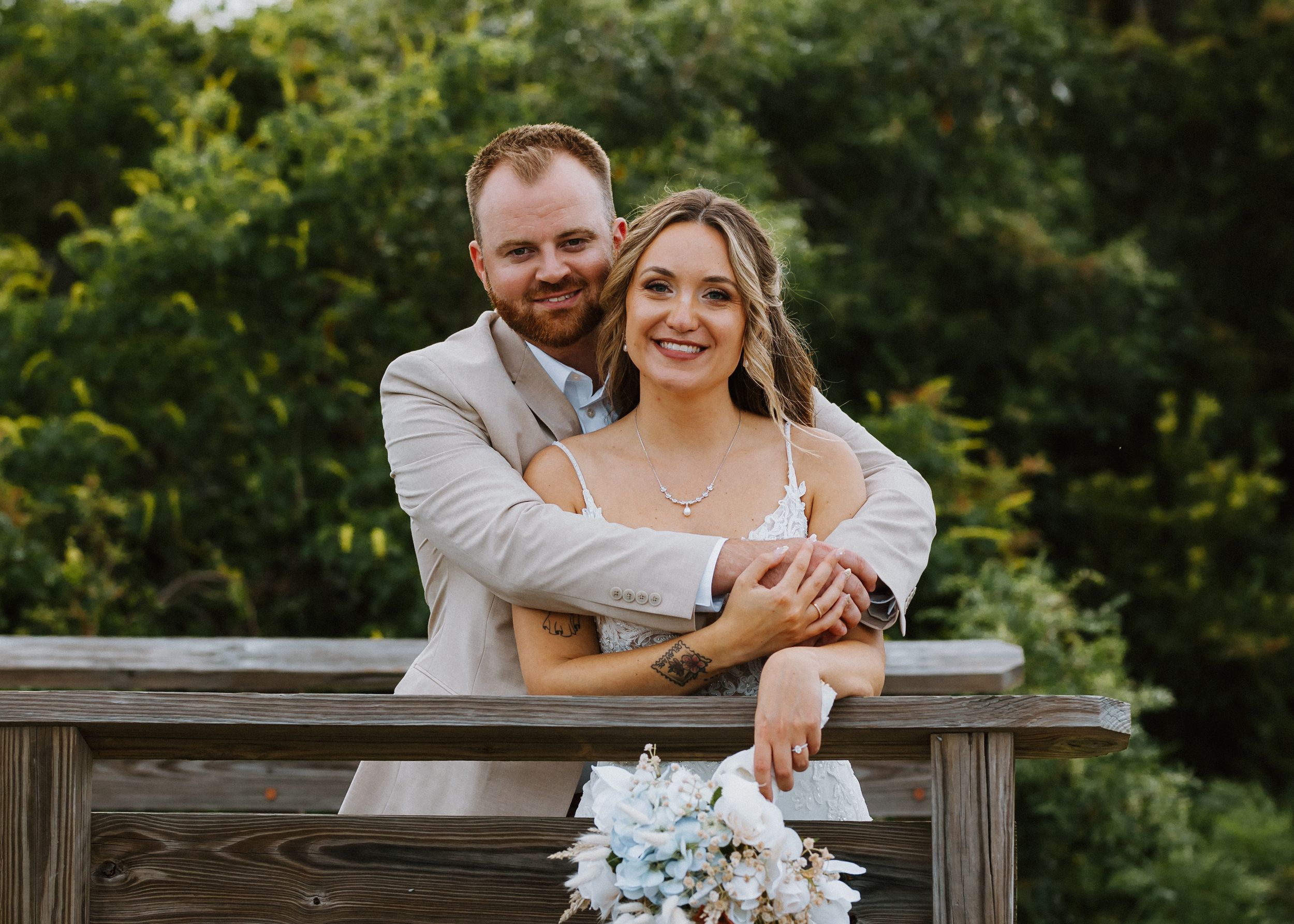 Smiling couple in wedding attire poses at wooden railing with blue floral bouquet outdoors.