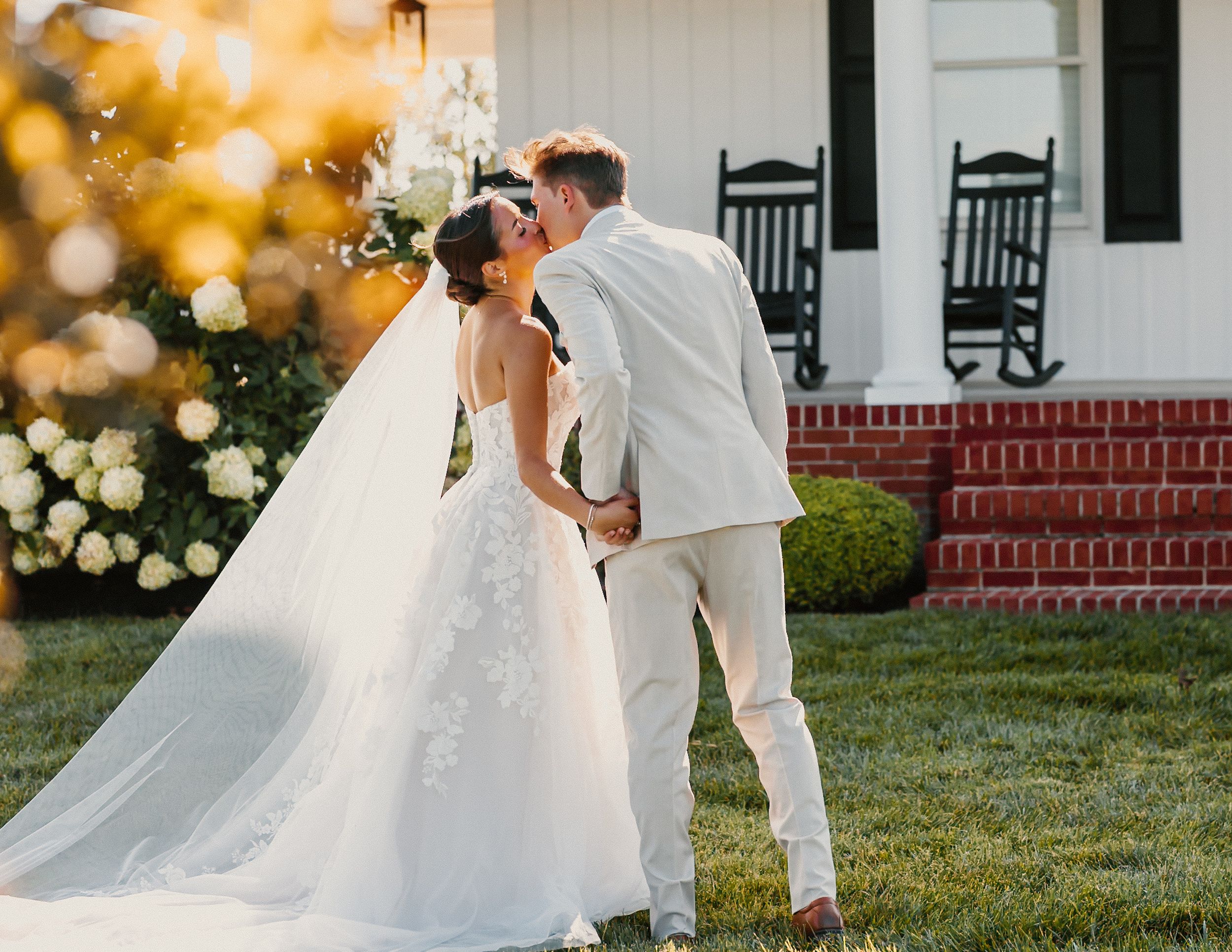 Bride and groom share a romantic kiss on a lawn outside a classic white farmhouse at golden hour.