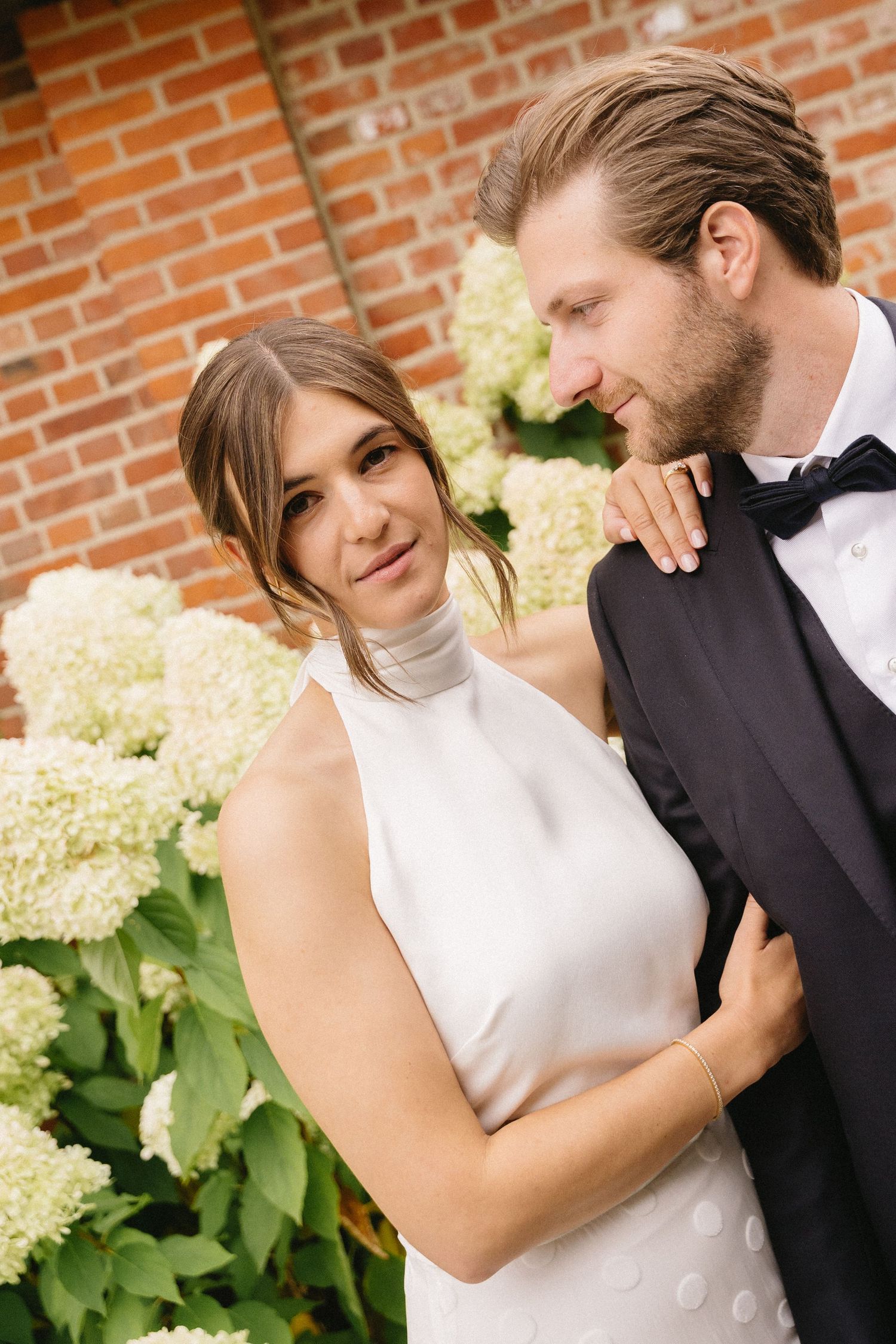 A tender moment between a couple in formal wedding attire against a brick wall with white hydrangeas.