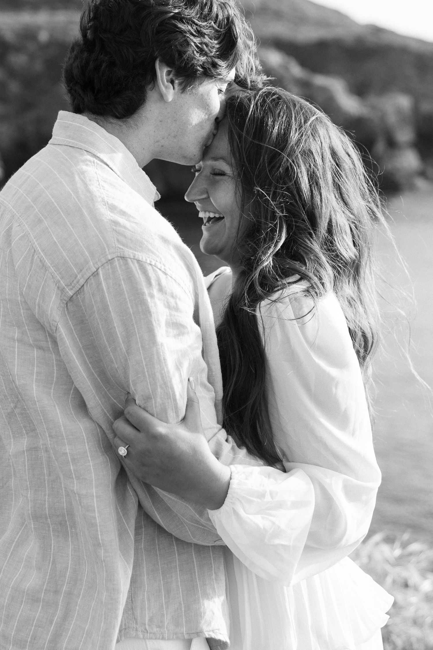 Joyful bride in white dress hugging groom in blue suit on beach.